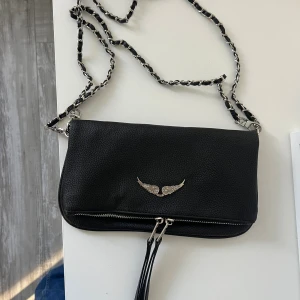 Svart axelväska med kedja och vingar - Svart axelväska i skinn med silverfärgad kedja och detaljer. Framsidan har ett metallmärke med vingar och en dubbel dragkedja med långa remmar. Baksidan har ytterfack med dragkedja. Stilren och edgy design, perfekt för dig som gillar coola accessoarer.oanvänd 