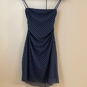 Randig marinblå klänning från Bershka - Säljer en marinblå kortklänning från Bershka med tunna axelband och vita diagonala ränder. Klänningen har en figurnära passform upptill och ett lätt utsvängt fall. Perfekt för dig som gillar en stilren och fräsch look.