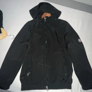 Svart vindjacka från Moncler med huva - Svart vindjacka från Moncler med huva, dragkedja framtill och två sidofickor med dragkedja. Jackan har Moncler-logga på ärmen och är tillverkad i lätt syntetmaterial. Perfekt för dig som gillar stilren och sportig look.
