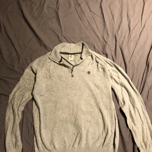 Grå Timberland half zip tröja - Snygg grå tröja från Timberland med halv dragkedja och ribbad krage. Tröjan har broderad logga på bröstet och är tillverkad i mjuk bomull. Perfekt för lager-på-lager och chill stil.