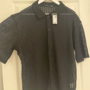 2 st nya Vit och svart stickad skjorta från 157 - Två skjortor från 157, en i svart och en i vit, båda i stickad ribbad design med korta ärmar och klassisk krage. Skjortorna har knappar framtill och är tillverkade i ett luftigt stickat material som ger en cool och modern vibe. Perfekt för dig som gillar att sticka ut med stil. Nya med tags storlek M