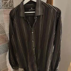 Randig skjorta från Batistini, L - Snygg skjorta från Batistini i storlek L med bruna, beige och svarta ränder. Klassisk krage och knappar framtill. Långärmad modell med smal passform och coolt mönster som ger en stilren vibe. Perfekt för dig som gillar detaljer och vill sticka ut lite extra.