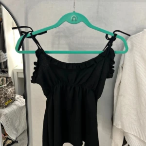 Svart blus med rosett från 157 - Supersöt svart blus från 157 med knytband på axlarna och volangdetaljer längs halsringningen. Blusen har en loose passform och är ärmlös, perfekt för dig som gillar en chill och trendig look. Materialet känns mjukt och lätt mot huden. Pris kan diskuteras 🥰