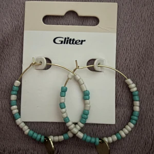 Hoops med pärlor från Glitter - Stora creolörhängen i guldmetall från Glitter, dekorerade med turkosa och vita pärlor samt en liten guldig berlock längst ner.