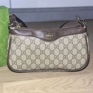 Väska - En helt ny väska som aldrig är använd. Väskan kommer med en extra förlängning, så att bandet på väskan blir längre. När man köper så följer även boxen med. Om du har några frågor eller vill ha fler bilder så är det bara att fråga.  