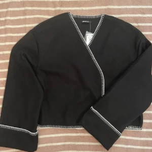 Svart omlottkofta med vita sömmar - Svart omlottkofta från Gina Tricot med kontrasterande vita dekorationssömmar längs kanterna. Koftan har v-ringning, vida ärmar och croppad passform. Perfekt för lager-på-lager och ger en stilren men ändå chill vibe.