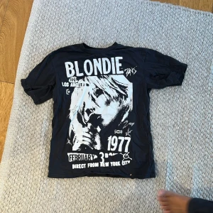 Svart Blondie t-shirt från H&M - Cool svart t-shirt från H&M med stort Blondie-tryck i vitt på framsidan. T-shirten har rund hals, korta ärmar och är gjord i mjuk bomull. Trycket har en retro känsla med text och bild från Blondies spelning 1977. Perfekt för dig som gillar bandtröjor och streetstyle.
