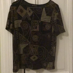 Svart topp med geometriskt mönster Soft Works - Cool svart topp från Soft Works med korta ärmar och ett unikt geometriskt mönster i guld och silver. Mönstret består av spiraler, trianglar och linjer som ger en riktigt artsy vibe. Materialet har en lätt glittrig finish som syns snyggt i ljuset. Perfekt för dig som gillar att sticka ut.