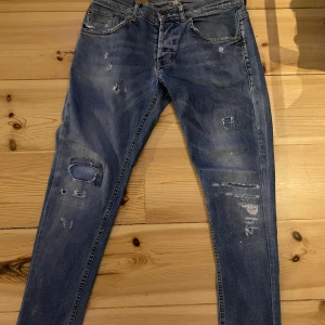 Blå jeans med slitningar från Dondup - Snygga blå jeans från Dondup med slitna detaljer och lagningar. Jeansen har knappar med Dondup-logga och en metall-D på bakfickan. Perfekt för dig som gillar en cool och avslappnad stil. Priset är absolut inte slaget i sten så var inte rädd att ge ett bud.