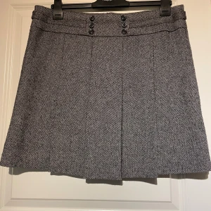 Grå kortkjol med knappar - Snygg grå kortkjol från Esprit, med subtilt mönster och dekorativa knappar framtill i midjan. Kjolen har pressveck som ger en stilren och klassisk look. Perfekt att matcha med en stickad tröja eller skjorta. Kjolennär helfodrad. Storlek 42