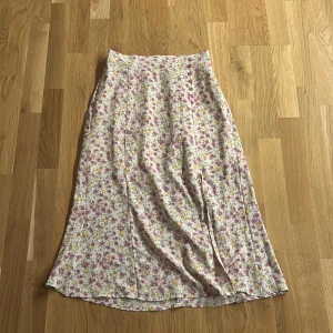 Blommig långkjol med knappar - Supersöt mellanlångkjol med blommigt mönster. Kjolen har en rad lila knappar framtill och den har även en split vid vänstra benet