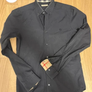 Svart skjorta från Burberry - Stilren svart skjorta från Burberry med klassisk krage och knappar framtill. Ikoniskt rutigt tyg syns på insidan av manschetterna och kragen. Broderad logga på bröstet ger en exklusiv touch. Perfekt för dig som gillar snygga detaljer och tidlös design.
