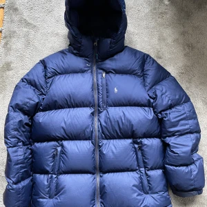 Mörkblå pufferjacka (dunjacka) från Polo Ralph Lauren - Säljer en mörkblå pufferjacka (dunjacka) från Polo Ralph Lauren Performance med huva och två sidofickor med dragkedja. Jackan har en klassisk Polo-logga på bröstet. Perfekt för kalla dagar med sin tjocka och quiltade design.