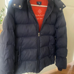 Mörkblå pufferjacka från GANT - Mörkblå vinterjacka från GANT strl 158-164( 13-14 år) med orange foder. Jackan har huva med tryckknappar, två sidofickor och dragkedja framtill. Ett litet hål har lagats med en patch, syns knappt vid användning. Annars inga skador eller märken.