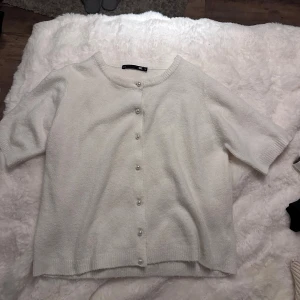 Vit stickad kofta med pärlknappar - Superfin vit stickad kofta från lager 157 med korta ärmar och rund hals. Framsidan har dekorativa pärlknappar som ger en söt touch. Perfekt att slänga över en topp för en chill look. Mjuk och skön kvalitet, passar dig som gillar enkel och stilren design.
