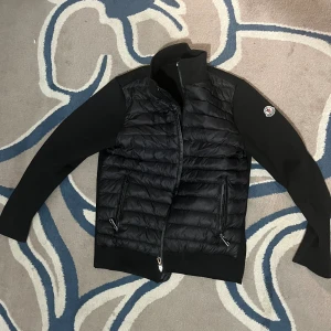 Moncler cardigan jacka - Svart Moncler storlek S med quiltad front och släta ärmar. Jackan har ståkrage, två dragkedjeförsedda fickor och Moncler-logga på ena ärmen. Materialmix med polyester och stickade detaljer. Perfekt för dig som gillar stilren och sportig look.