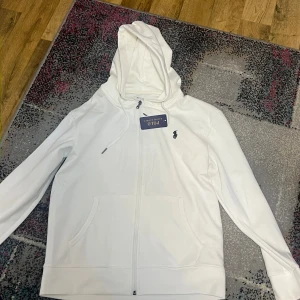 Vit hoodie från Polo Ralph Lauren - Vit hoodie från Polo Ralph Lauren med dragkedja framtill, huva med snörning och klassisk logga på bröstet. Tröjan har känguruficka och är tillverkad i mjuk bomull. Perfekt för en clean och stilren look.