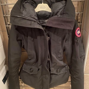  Canada Goose Montebello parka XS - Svart dunjacka från Canada Goose med stor huva, dragkedja och knappar framtill. Jackan har två stora fickor med lock och knapp, samt en ärmficka med dragkedja. Klassisk röd och vit Canada Goose-patch på ärmen. Perfekt för kalla vinterdagar.