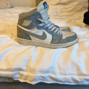 Nike Air Jordan 1 High blå/vit - Nike Air Jordan 1 High sneakers i blått och vitt med klassisk Jordan-logga på sidan. Skorna har snörning, perforerad tå och platt sula. Materialet är en mix av skinn och mocka, med blå detaljer och vit bas. Perfekt för dig som gillar retrostil och streetwear.