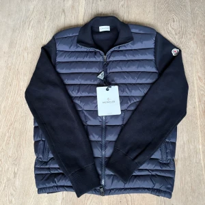 Moncler cardigan - Säljer en svart pufferjacka från Moncler med quiltad front i polyester och stickade ärmar. Jackan har Moncler-logga på ärmen, ribbad krage och dragkedja framtill. Snygg och stilren design som passar perfekt till streetwear och chill vardagsstil. Aldrig andvänd. Sitter lite mer som S men kan även passa M. Priset är ej hugget i sten.
