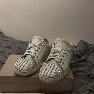Christian Louboutin sneakers med nitar - Säljer ett par vita sneakers från Christian Louboutin med ikoniska röda sulor och silverfärgade nitar över tå och häl. Skorna har snörning och är tillverkade i skinn med en cool och edgy vibe. Perfekta för dig som vill sticka ut med din stil.