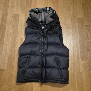 Svart väst från Burberry - Stilren svart väst från Burberry med avtagbar huva och klassiskt rutigt foder i grått, svart och rött. Västen har dragkedja och tryckknappar framtill samt två snedställda fickor med dragkedja och en innerficka med dragkedja. Perfekt för kyliga dagar. En reva i insidan och ena dragsko snöret har lossnat på ena sidan men går att trycka in den. Inga andra skador. Bra skick