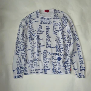Vit stickad Supreme-tröja med blå text - Supreme stickad tröja i vitt med blå handskrivna texter. Långärmad modell med rund hals och ribbade muddar. Tröjan har en unik streetstyle-vibe och är perfekt för dig som vill sticka ut.