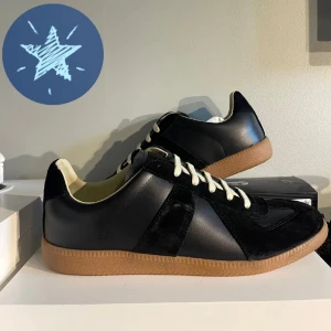 Maison Margiela skor - Maison Margiela sneakers i svart skinn och mocka med vita snören och beige innersula. Klassisk siluett med gummisula i brunt och snygga kontraster mellan material. Perfekta för dig som gillar stilrena och tidlösa sneakers med premiumkänsla.