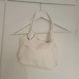 Vit axelväska från Mohito Accessories - Vit axelväska från Mohito Accessories. Väskan har en mjuk, slät yta, dubbla handtag och ett innerfack med dragkedja och magnetknapp