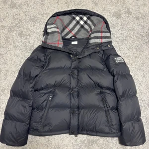 Svart Burberry pufferjacka med huva - Svart pufferjacka från Burberry med stor huva och klassiskt rutigt foder i grått, vitt och rött. Jackan har Burberry London England-logga på ärmen, dragkedjor på fickorna och knappar framtill. Perfekt för kalla dagar med sin dunfyllda design och snygga detaljer.