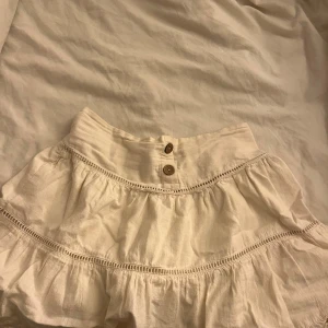 Vit volangkjol från Zara - Supersöt vit kortkjol från Zara med volanger och dekorativa knappar framtill. Kjolen har bred resår i midjan och fina spetsdetaljer längs volangerna. Perfekt för en fräsch och somrig look.