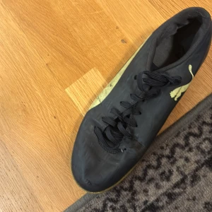 Svarta fotbollsskor från Puma - Svarta fotbollsskor från Puma med guldfärgad logga på sidan och snörning framtill. Skorna har en smal och sportig form, perfekt för dig som vill ha bra grepp på planen. Yttermaterialet är syntet och insidan är vadderad för extra komfort. Daha Ali har blivit tacklad i dem 