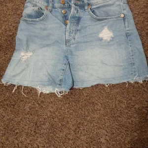 Blå denimshorts med slitningar 157 - Säljer ett par ljusblå denimshorts från 157 i storlek S. Shortsen har hög midja, råa och fransiga kanter samt slitna detaljer framtill. Klassisk femficksmodell med knappgylf och bälteshällor. Perfekta för en chill sommarlook.