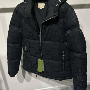 Svart pufferjacka från Gucci - Säljer en svart pufferjacka från Gucci med GG-logga över hela jackan. Jackan har huva med snörning, två fickor med dragkedja och en snygg, modern look. Perfekt för dig som vill ha en lyxig och trendig jacka i vinter.