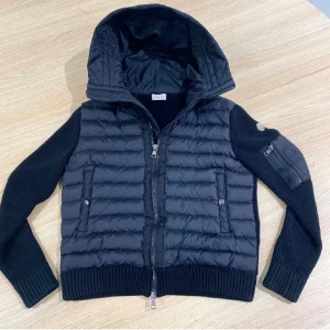 Svart jacka från Moncler - Svart pufferjacka från Moncler med stickade ärmar. Jackan har ribbade muddar, två sidofickor med knapp, dragkedja framtill och Moncler-logga på ärmen. Materialmix av polyester och stickad ull ger en schysst look och skön känsla.(säljer för köpte en ny jacka)