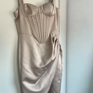Champagnefärgad bustierklänning - Supersnygg kort klänning i champagnefärgat satinliknande material med markerad bustier, draperad detalj vid höften och breda axelband. Figurnära passform och coola sömmar framtill ger en lyxig och trendig vibe.