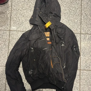 Svart bomberjacka från Parajumpers - Svart bomberjacka från Parajumpers med huva, flera fickor med dragkedja och coola detaljer som orange rem och broderade märken. Jackan har ribbad mudd vid midja och ärmar, samt slitstarka material och snygga kontrastsömmar. Perfekt för dig som gillar streetwear och funktion.