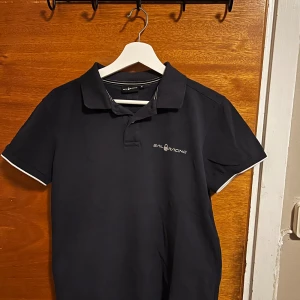 Sail Racing Mörklblå piké  - Svart pikétröja från Sail Racing med vit logga på bröstet och vit detalj på ärmsluten. Klassisk krage och korta ärmar, perfekt för en clean och sportig look. 