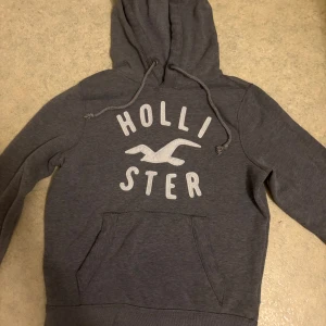 Grå hoodie från Hollister med tryck - Snygg grå hoodie från Hollister med stort vitt tryck och logga på bröstet. Tröjan har huva med snörning, magficka och långa ärmar. Perfekt för chill dagar och enkel att matcha med jeans eller joggers. Ända defekten med varan är att den är lite urtvättad.