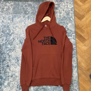 Brun hoodie North Face - Säljer en snygg rostbrun hoodie från The North Face med stor svart logga på bröstet. Tröjan har huva med snörning, magficka och långa ärmar. Perfekt för chill dagar och streetwear. Materialet är mjukt och känns som bomull och polyester. Pris kan diskuteras vid snabb affär