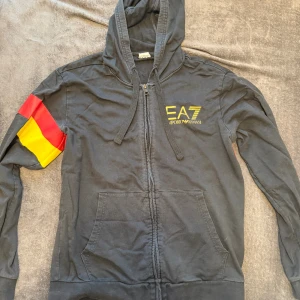 Svart EA7 hoodie med färgglada detaljer - Svart hoodie från EA7 Emporio Armani med dragkedja, huva och känguruficka. Snygga gula och röda detaljer på ärm och rygg samt stort EA7-tryck i gult framtill. Perfekt för dig som gillar streetwear och vill sticka ut.