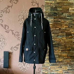 Svart parkajacka från RVLT - Svart fodrad  parkajacka från Revolution med vit snörning i huvan, stora fickor med knappar och dragkedja framtill. Jackan har lång ärm, justerbara muddar och en stilren look. 