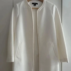 Vit kappa från H&M i strukturerad tyg - Säljer en stilren vit kappa från H&M med rak passform och diskret struktur i tyget. Jackan har lång ärm, två fickor fram och är helt utan knappar eller dragkedja för en clean look