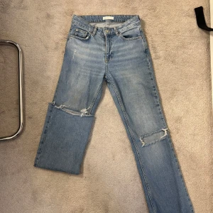 Ljusblå slitna jeans med hål - Säljer ett par ljusblå jeans med snygga slitningar. Klassisk femficksmodell med dragkedja och knapp. Jeansen har raka ben och en avslappnad vibe, perfekta för en cool streetstil.