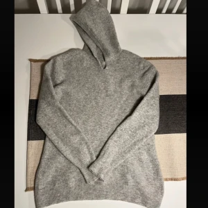 Grå hoodie i ullmix med huva 100% Kashmir - Mysig grå hoodie i mjuk ullmix med huva och långa ärmar. Tröjan har en enkel och stilren design med ribbade muddar vid ärmslut och nederkant. Perfekt för kyliga dagar när du vill vara både bekväm och snygg.