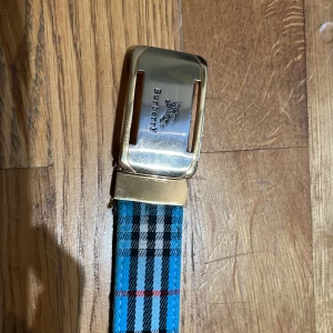 Blårutig Burberry bälte med guldspänne - Snyggt bälte från Burberry med klassiskt rutigt mönster i blått, svart, vitt och rött. Bältet har ett stilrent guldigt metallspänne med Burberry-logga ingraverad. Perfekt för att lyfta din outfit med en exklusiv touch.