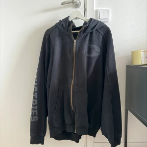 Gasp hoodie - Svart hoodie från Gasp  med huva, dragkedja framtill och stora fickor. Tryck på ena ärmen och broderad logga på bröstet. Skön passform och ribbade muddar vid ärmslut och nederkant. Perfekt för chill dagar.