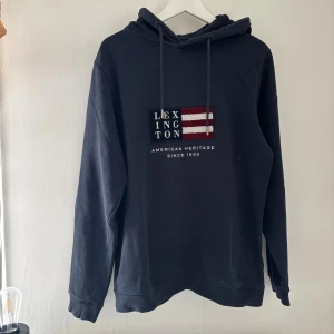 Mörkblå hoodie från Lexington - Snygg mörkblå hoodie från Lexington med broderad amerikansk flagga och text på bröstet. Tröjan har huva med snörning och långa ärmar. Perfekt för chill dagar och enkel att matcha med jeans eller joggers.