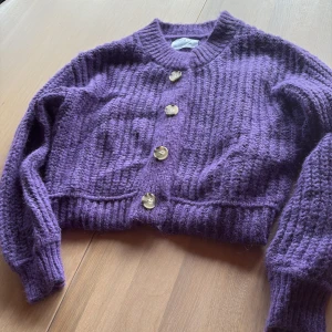 Lila stickad kofta från Zara - Säljer en lila stickad kofta från Zara med grov ribbad struktur och fyra stora knappar framtill. Koftan har rund hals, långa ärmar och är croppad i modellen. Perfekt för dig som gillar färg och vill ha en mysig tröja med cool vintagekänsla.