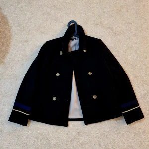 Marinblå kappa med guldknappar - Stilren marinblå kappa med dubbla rader guldknappar framtill, broderad text 'Lovers' i rött på ryggen och coola detaljer som guldstjärnor och blå/guldfärgade band på ärmarna. Kappan hög krage och är helfodrad för extra komfort.